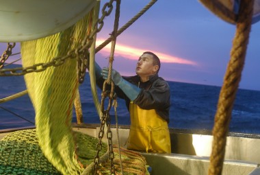 Marin pêcheur déroulant une partie du chalut en vue de sa réparation, sur fond de lever de soleil.